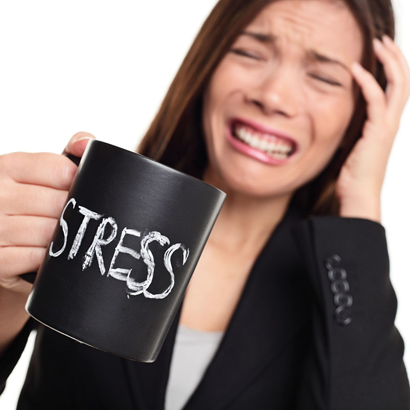 woman grimacing with one hand to her head and the other holding a mug with the word stress on it.